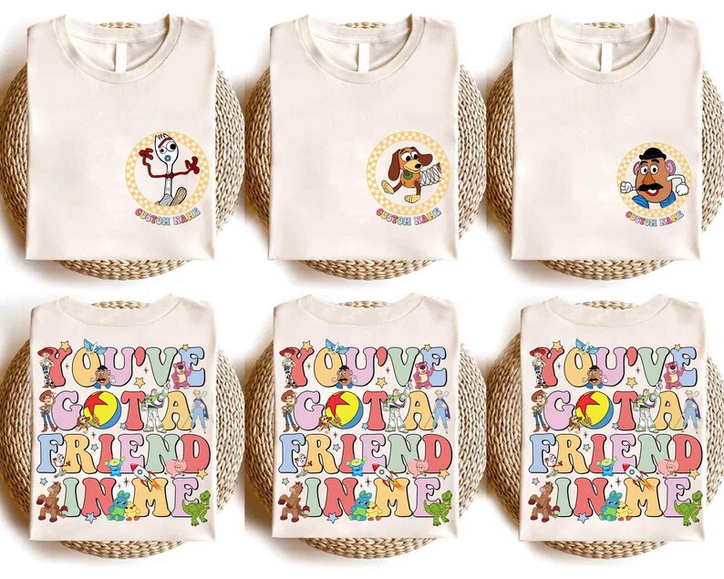 Op de afbeelding: Zes cr&egrave;mekleurige t-shirts met kleurrijke graphics. De bovenste rij heeft karakterontwerpen in cirkels, terwijl de onderste rij de tekst "YOU'VE GOT A FRIEND IN ME" in een retro lettertype weergeeft, met verschillende karakterillustraties.