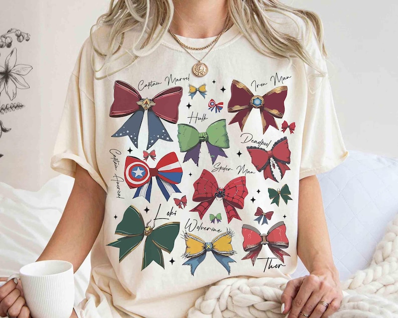 May include: Cream-colored t-shirt featuring colorful bow designs representing Marvel characters like Captain Marvel, Iron Man, and Thor. Each bow is uniquely colored and labeled with the character's name in a handwritten font. The shirt is worn by a person.