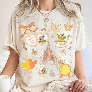 May include: Cream-colored t-shirt featuring a collection of orange-themed illustrations. Designs include a castle, orange slices, Mickey Mouse balloons, and a heart-shaped graphic with the text "Sunshine On my mind". The shirt has a relaxed fit.