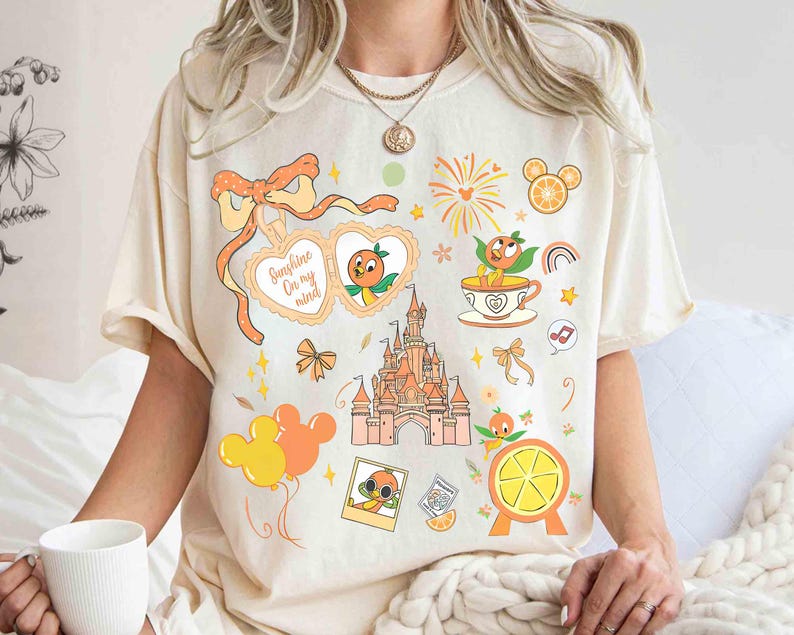 May include: Cream-colored t-shirt featuring a collection of orange-themed illustrations. Designs include a castle, orange slices, Mickey Mouse balloons, and a heart-shaped graphic with the text "Sunshine On my mind". The shirt has a relaxed fit.