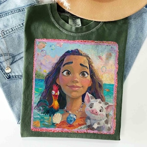 Puede incluir: Una camiseta de cuello redondo color verde oliva con un estampado gr&aacute;fico colorido y pict&oacute;rico de una joven de pelo largo y oscuro, un gallo y un cerdito. El dise&ntilde;o se sit&uacute;a sobre un fondo de oc&eacute;ano tropical con burbujas y un borde rosa.