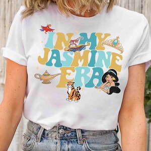 Op de afbeelding: Wit T-shirt met de tekst "IN MY JASMINE ERA" in turquoise en gouden letters, met cartoon illustraties van een papegaai, vliegend tapijt, kroon, lamp, tijger en prinses Jasmine. Het shirt heeft een ronde hals en korte mouwen.