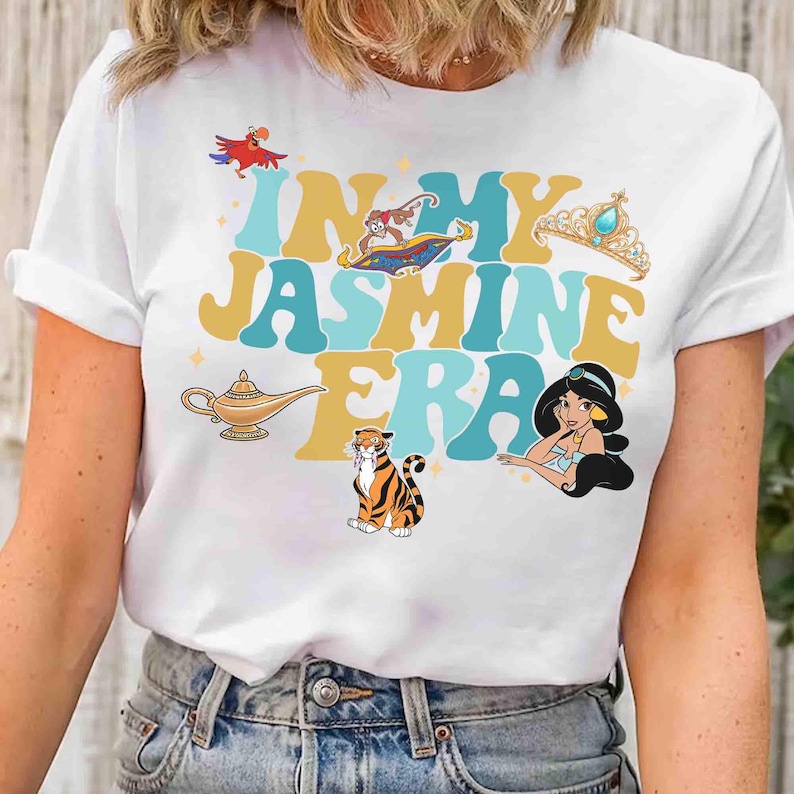 Op de afbeelding: Wit T-shirt met de tekst "IN MY JASMINE ERA" in turquoise en gouden letters, met cartoon illustraties van een papegaai, vliegend tapijt, kroon, lamp, tijger en prinses Jasmine. Het shirt heeft een ronde hals en korte mouwen.