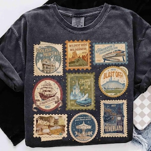 以下が含まれることがあります： チャコールグレーのTシャツで、9枚のヴィンテージ風切手のグラフィックプリントが施されています。各切手には、「Foolish Mortals」、「Wildest Ride in the Wilderness」、「Tomorrow's Transportation」、「A Pirates Life」、「Happily Ever After」、「Blast Off」、「Heigh-Ho」、メリーゴーランド、「Off to Neverland」といったユニークなイラストとテキストが描かれています。