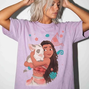 Puede incluir: Una camiseta de manga corta de color morado claro con un estampado gr&aacute;fico de un personaje de pelo largo abrazando a un peque&ntilde;o cerdito. El dise&ntilde;o incluye peque&ntilde;os iconos coloridos de flores y hojas, adem&aacute;s de una vela estilizada.