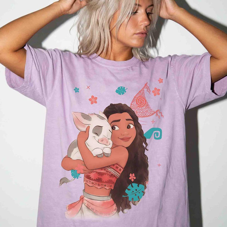 Puede incluir: Una camiseta de manga corta de color morado claro con un estampado gr&aacute;fico de un personaje de pelo largo abrazando a un peque&ntilde;o cerdito. El dise&ntilde;o incluye peque&ntilde;os iconos coloridos de flores y hojas, adem&aacute;s de una vela estilizada.