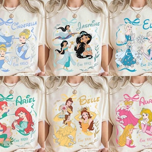 K&ouml;nnte beinhalten: Cremefarbene T-Shirts mit verschiedenen Disney-Prinzessinnen: Cinderella, Jasmin, Elsa, Arielle, Belle und Aurora. Jedes Shirt zeigt den Namen der Prinzessin, Illustrationen und das Jahr ihres Deb&uuml;ts. Die Designs sind mit dekorativen Elementen umrahmt.