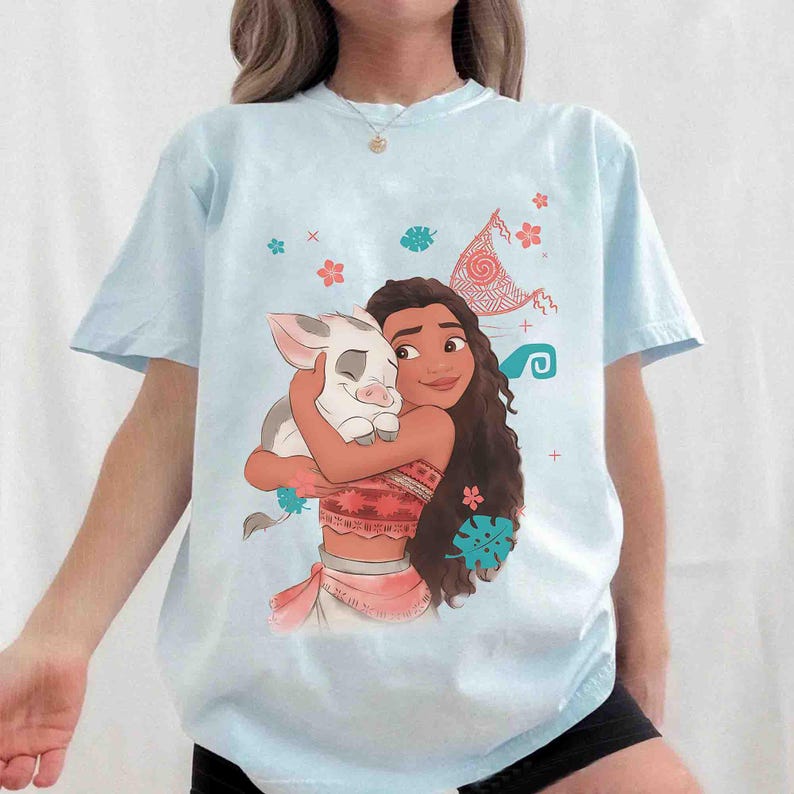 Puede incluir: Una camiseta de manga corta azul claro con una ilustraci&oacute;n gr&aacute;fica de una persona joven de pelo largo, oscuro y ondulado abrazando a un peque&ntilde;o cerdo blanco. El dise&ntilde;o incluye detalles florales, una forma de vela y peque&ntilde;os s&iacute;mbolos.
