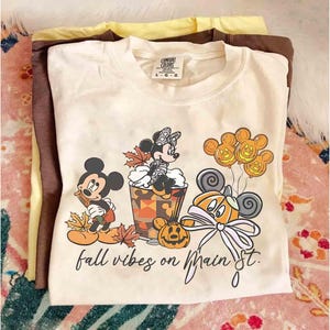 Pode incluir: Camiseta Comfort Colors creme com Mickey e Minnie Mouse em trajes de outono. O gráfico inclui abóboras, folhas de outono e o texto "fall vibes on Main St.". A camisa é dobrada com outras camisas.