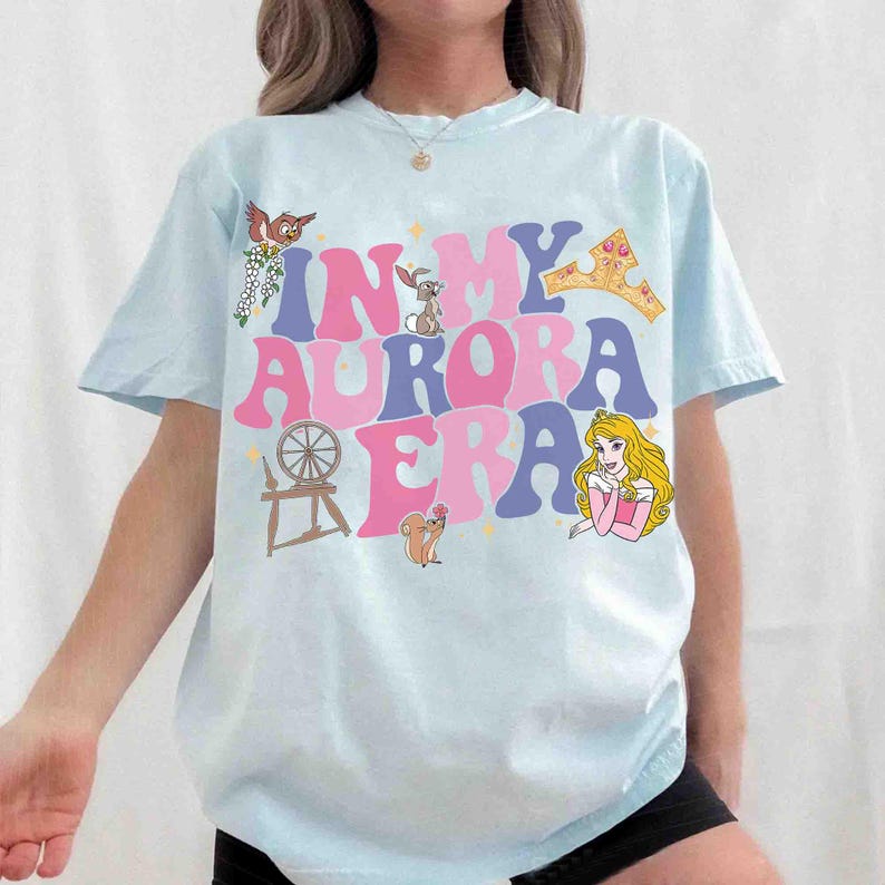 Op de afbeelding: Lichtblauw T-shirt met de tekst "IN MY AURORA ERA" in roze en blauwe letters. Het shirt heeft cartoon illustraties van een vogel, konijn, kroon, spinnewiel, eekhoorn en een prinses. Het shirt heeft korte mouwen.