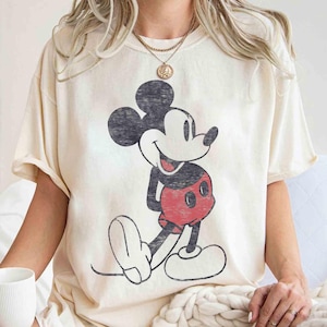 Könnte beinhalten: Cremefarbenes T-Shirt mit einem verblichenen Mickey Mouse-Motiv. Die Zeichentrickfigur ist in Schwarz, Rot und Weiß dargestellt. Das Shirt hat eine lockere Passform und kurze Ärmel, perfekt für den Alltag.
