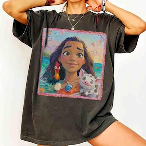 Puede incluir: Una camiseta de manga corta gris carb&oacute;n con un estampado gr&aacute;fico cuadrado de una persona joven con cabello largo y oscuro, un gallo y un cerdo peque&ntilde;o. La escena se sit&uacute;a sobre un fondo de oc&eacute;ano azul con burbujas, enmarcada por un borde rosa.