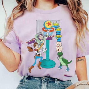 May include: A light purple t-shirt featuring a basketball hoop with cartoon characters Buzz Lightyear, Woody, and other Toy Story characters. The design includes a basketball, aliens, and a dinosaur.