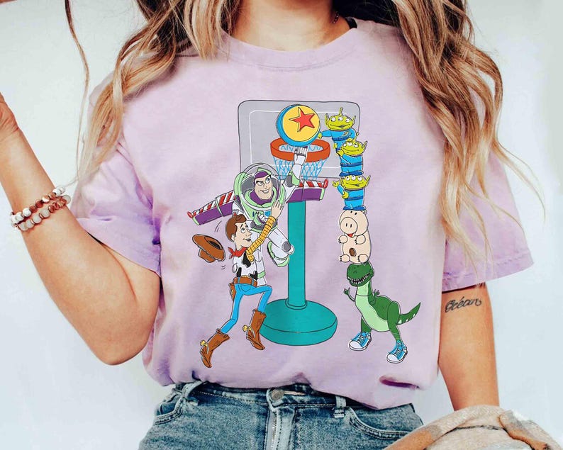 May include: A light purple t-shirt featuring a basketball hoop with cartoon characters Buzz Lightyear, Woody, and other Toy Story characters. The design includes a basketball, aliens, and a dinosaur.