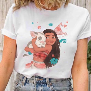 Puede incluir: Una camiseta blanca de manga corta con una ilustraci&oacute;n gr&aacute;fica de una persona joven de pelo largo y casta&ntilde;o abrazando a un peque&ntilde;o lech&oacute;n. El dise&ntilde;o incluye elementos decorativos como flores rojas, hojas verde azulado y una vela.