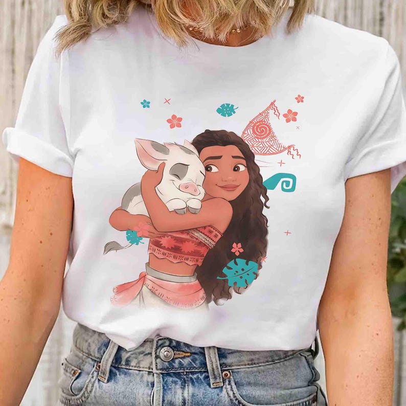 Puede incluir: Una camiseta blanca de manga corta con una ilustraci&oacute;n gr&aacute;fica de una persona joven de pelo largo y casta&ntilde;o abrazando a un peque&ntilde;o lech&oacute;n. El dise&ntilde;o incluye elementos decorativos como flores rojas, hojas verde azulado y una vela.