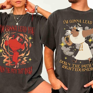 May include: Two dark gray t-shirts with cartoon graphics. One shirt features a red devil with the text "I'M GONNA LEAD YOU DOWN THE PATH THAT ROCKS." The other shirt shows a character in white with the text "I'M GONNA LEAD YOU DOWN THE PATH OF RIGHTEOUSNESS."