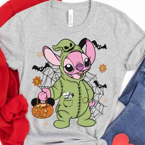 May include: A light gray t-shirt featuring a cartoon character in a green costume holding a pumpkin-shaped basket. The character has pink ears and is surrounded by bats, spiderwebs, and flowers. The shirt is paired with blue jeans.