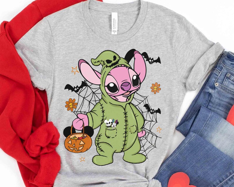 May include: A light gray t-shirt featuring a cartoon character in a green costume holding a pumpkin-shaped basket. The character has pink ears and is surrounded by bats, spiderwebs, and flowers. The shirt is paired with blue jeans.