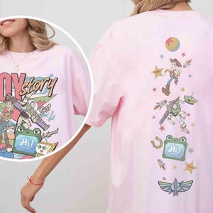 Op de afbeelding: Een lichtroze t-shirt met een Toy Story-afbeelding. De print op de voorkant toont de personages Jessie, Woody, Buzz en anderen met de tekst "TOY story" en "SINCE 1995". De achterkant heeft een verticale opstelling van personages, sterren en een "Hi!"-bord.