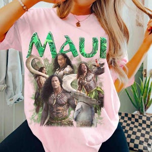 May include: A light pink t-shirt featuring a large graphic print of the character Maui from the movie Moana. The design includes the text "MAUI" in bold green letters, multiple depictions of the character, a shark, and green lightning accents.