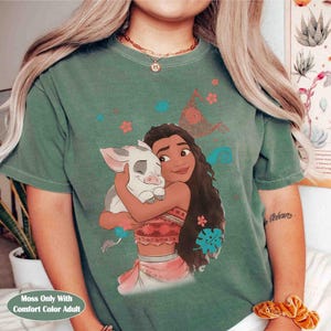 Puede incluir: Una camiseta de manga corta color verde salvia con un estampado gr&aacute;fico de un personaje de pelo largo abrazando a un peque&ntilde;o cerdo blanco. El dise&ntilde;o incluye elementos florales dispersos en rojo, azul y naranja, junto a un patr&oacute;n de espiral.
