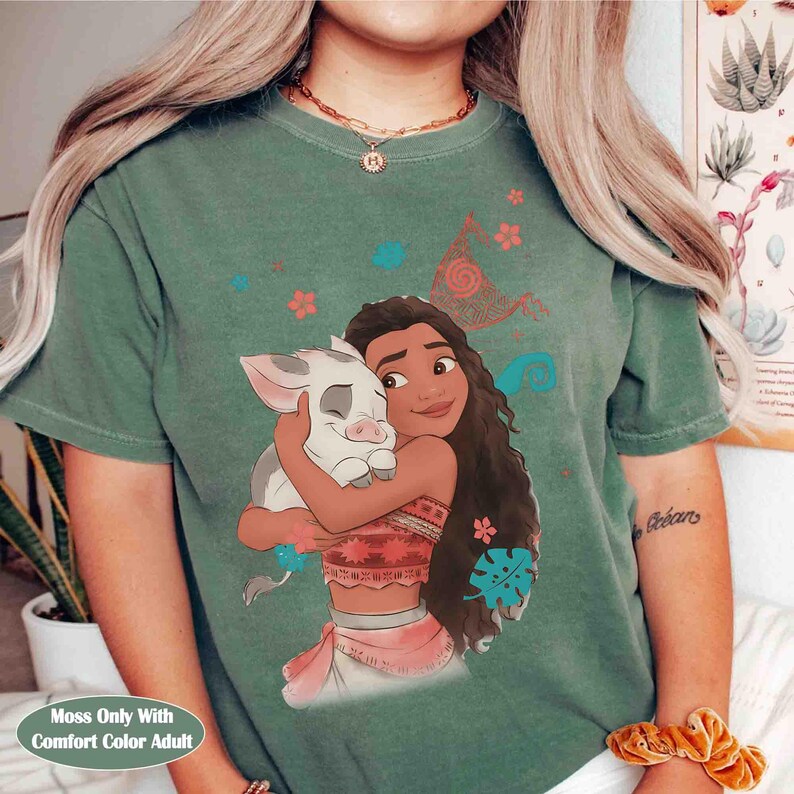 Puede incluir: Una camiseta de manga corta color verde salvia con un estampado gr&aacute;fico de un personaje de pelo largo abrazando a un peque&ntilde;o cerdo blanco. El dise&ntilde;o incluye elementos florales dispersos en rojo, azul y naranja, junto a un patr&oacute;n de espiral.