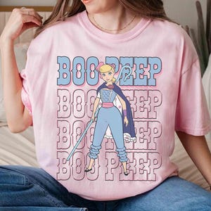 May include: A light pink t-shirt featuring a cartoon graphic of Bo Peep from Toy Story. The graphic includes the text "BOO PEEP" in blue and pink, with Bo Peep holding a staff and wearing a cape and blue outfit.