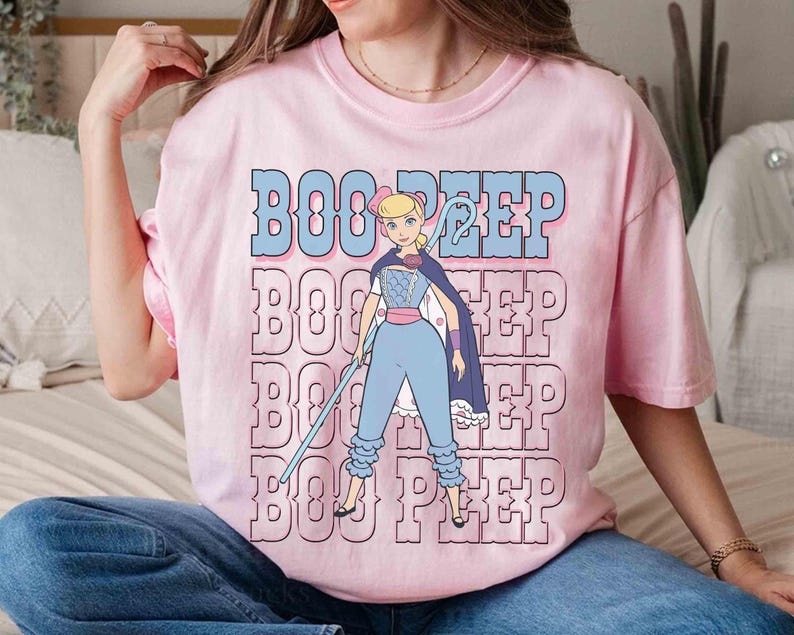 May include: A light pink t-shirt featuring a cartoon graphic of Bo Peep from Toy Story. The graphic includes the text "BOO PEEP" in blue and pink, with Bo Peep holding a staff and wearing a cape and blue outfit.