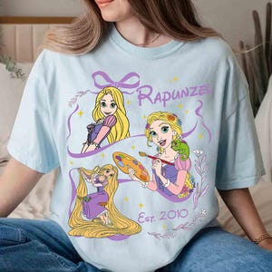 K&ouml;nnte beinhalten: Hellblaues T-Shirt mit drei Illustrationen von Rapunzel mit langen blonden Haaren, die lila und rosa Kleider tr&auml;gt. Das Wort "Rapunzel" steht &uuml;ber den Illustrationen, und "Est. 2010" steht darunter. Das Shirt ist aus weichem Material.