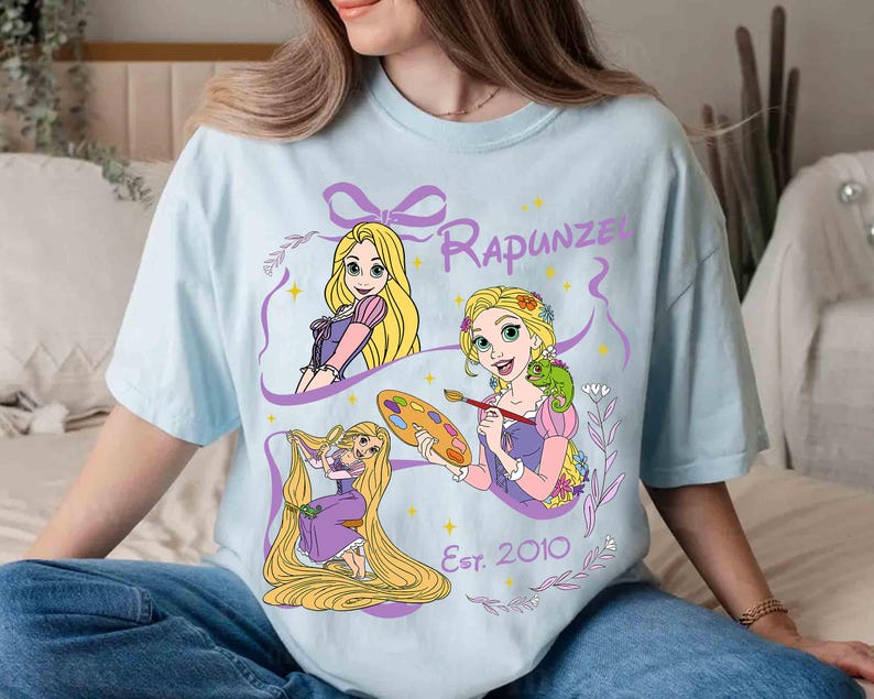 K&ouml;nnte beinhalten: Hellblaues T-Shirt mit drei Illustrationen von Rapunzel mit langen blonden Haaren, die lila und rosa Kleider tr&auml;gt. Das Wort "Rapunzel" steht &uuml;ber den Illustrationen, und "Est. 2010" steht darunter. Das Shirt ist aus weichem Material.