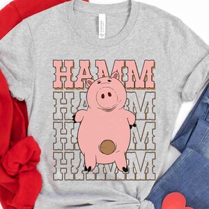 May include: A light gray t-shirt featuring a cartoon image of Hamm, the pink piggy bank character. The word "HAMM" is printed above the pig in a pink, retro-style font. The shirt is made of a soft material.