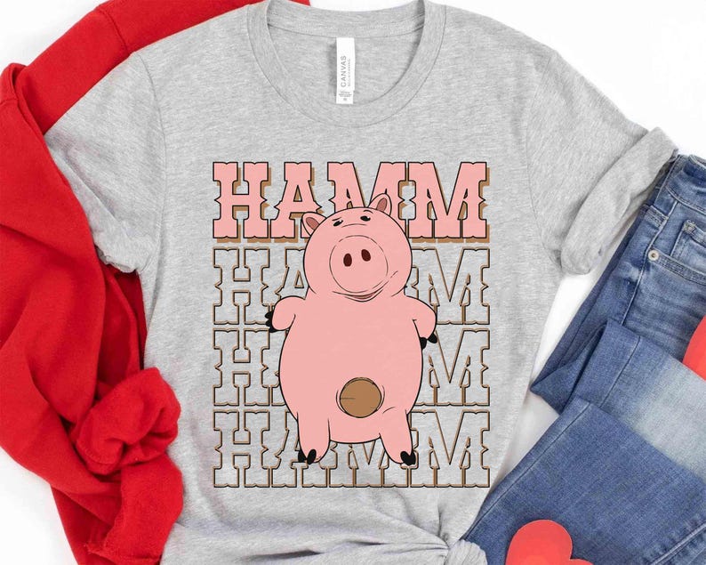 May include: A light gray t-shirt featuring a cartoon image of Hamm, the pink piggy bank character. The word "HAMM" is printed above the pig in a pink, retro-style font. The shirt is made of a soft material.