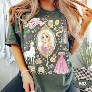 May include: Moss green t-shirt featuring a cartoon illustration of a princess in a mirror, surrounded by related imagery. Includes a white horse, pink dress, guitar, and the text "and at last I see the light".