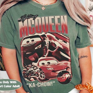 K&ouml;nnte beinhalten: Ein moosgr&uuml;nes T-Shirt mit einer Grafik von Lightning McQueen aus dem Film Cars. Das Design enth&auml;lt den Text "Lightning McQueen" und den Satz "KA-CHOW!". Das Shirt ist mit "Moss Only With Comfort Color Adult" gekennzeichnet.