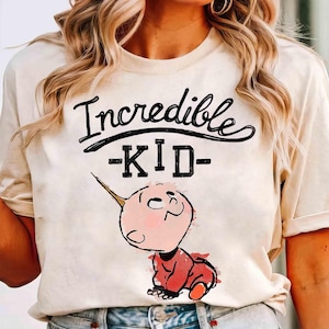 May include: Cream-colored t-shirt with the words "Incredible - KID -" in black script and block letters. The shirt features a cartoon illustration of a baby with a horn, wearing a red outfit.