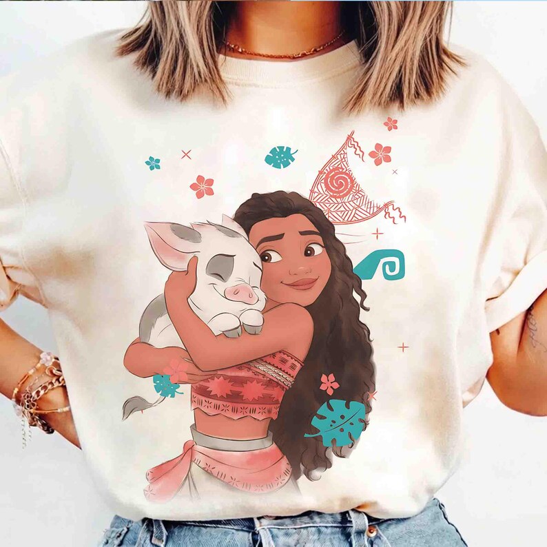 Puede incluir: Una camiseta de color crema con una ilustraci&oacute;n gr&aacute;fica de un personaje de pelo largo y casta&ntilde;o abrazando a un peque&ntilde;o lech&oacute;n blanco. El dise&ntilde;o incluye elementos decorativos como flores tropicales rojas y verde azulado, hojas y una vela.