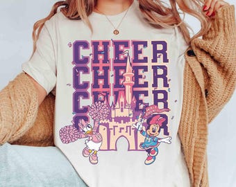 May include: Cream-colored t-shirt featuring the word "CHEER" in a purple and pink gradient, with a castle graphic and Disney characters Daisy Duck and Minnie Mouse. The shirt has a crew neck and short sleeves.