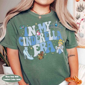 Op de afbeelding: Een mosgroen t-shirt met de tekst "IN MY CINDERELLA ERA" in wit en blauw. Het shirt heeft illustraties van Assepoester, een glazen muiltje, een kroon en andere gerelateerde afbeeldingen. Het shirt is gemaakt van een zacht, comfortabel materiaal.