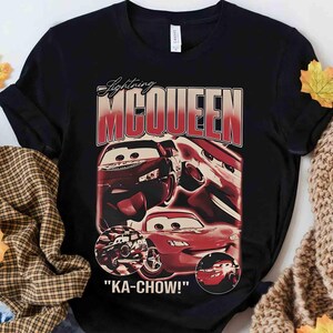 K&ouml;nnte beinhalten: Schwarzes T-Shirt mit einer Grafik von Lightning McQueen aus dem Film Cars. Das Design enth&auml;lt den Text "Lightning McQueen" in roter und wei&szlig;er Schrift sowie den Satz "KA-CHOW!" unter den Autobildern.