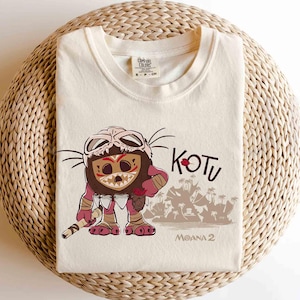 Puede incluir: Camiseta de color crema con un personaje de dibujos animados con una m&aacute;scara de calavera, sosteniendo un bast&oacute;n. La palabra "KOTU" est&aacute; encima del personaje, con "MOANA 2" debajo. La camiseta est&aacute; doblada sobre una superficie tejida.