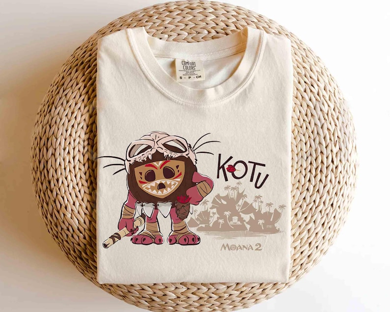 Puede incluir: Camiseta de color crema con un personaje de dibujos animados con una m&aacute;scara de calavera, sosteniendo un bast&oacute;n. La palabra "KOTU" est&aacute; encima del personaje, con "MOANA 2" debajo. La camiseta est&aacute; doblada sobre una superficie tejida.