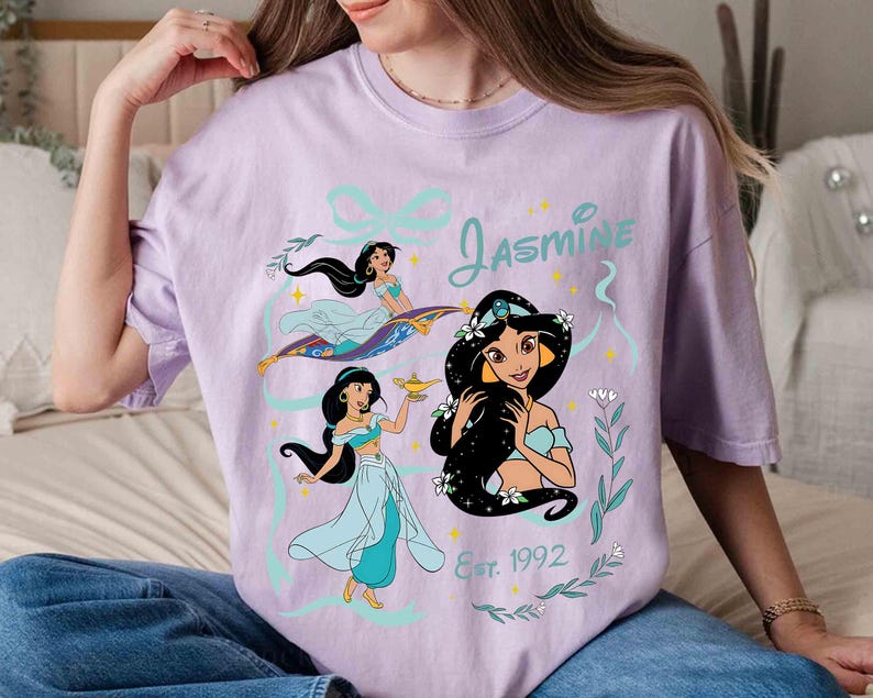 K&ouml;nnte beinhalten: Ein helllila T-Shirt mit einer Grafik von Prinzessin Jasmin aus Aladdin. Das Design umfasst drei Illustrationen von Jasmin, den Namen "Jasmine" in t&uuml;rkisfarbener Schrift und den Text "Est. 1992".