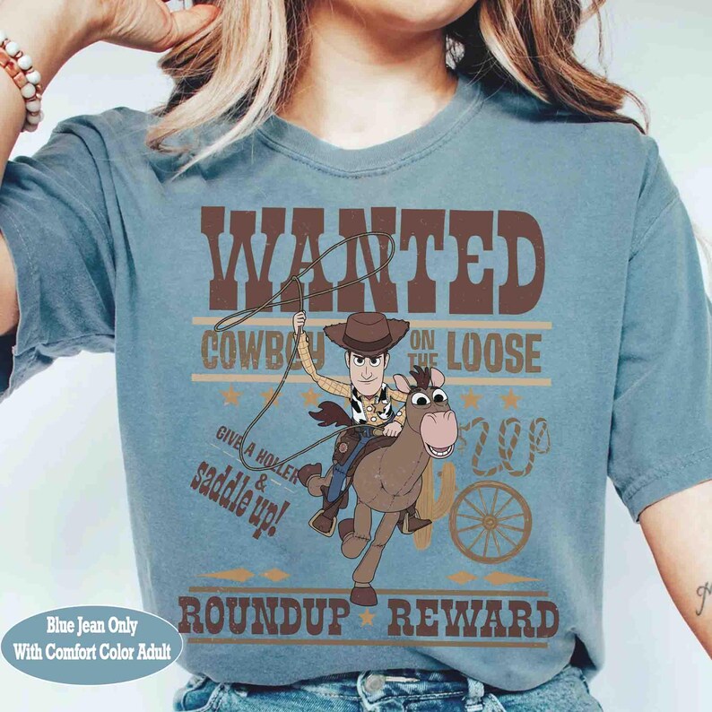 Pode incluir: Camiseta azul jeans confort&aacute;vel para adultos com um gr&aacute;fico de um cowboy montando um cavalo, com o texto "WANTED COWBOY ON THE LOOSE" e "ROUNDUP REWARD". A camisa tamb&eacute;m tem o texto "GIVE A HOLLER & SADDLE UP!"