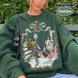 May include: A forest green sweatshirt featuring a graphic print of a green frog character wearing a scarf and top hat, standing near a castle in a snowy landscape. The design includes a decorative ribbon border and a small frog figure.