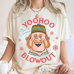 May include: Cream-colored t-shirt featuring a cartoon illustration of a smiling man with a red beard and a hat. The text "YOO HOO BLOWOUT" is printed in red, with "Big Summer" in blue script. Red snowflakes are scattered around the design.