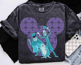 May include: A charcoal gray short-sleeve t-shirt featuring a large purple Mickey Mouse-shaped silhouette filled with a repeating wallpaper pattern. Three teal-colored ghost characters are printed in the center, wearing hats and formal attire.