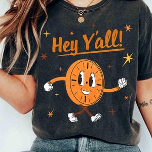 May include: Dark gray t-shirt with the text "Hey Y'all!" above a cartoon clock character. The clock is orange with white gloves and shoes, and is smiling. Gold stars and cross shapes surround the clock.