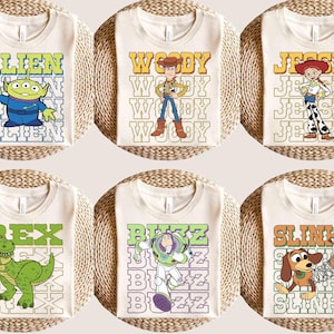 May include: Six cream-colored t-shirts featuring characters from a popular animated film. Each shirt displays a different character: Alien, Woody, Jessie, Rex, Buzz, and Slinky, with their names in a retro font. The shirts are arranged on a woven surface.