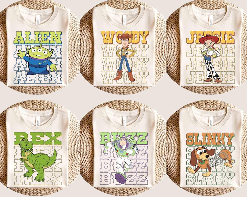 May include: Six cream-colored t-shirts featuring characters from a popular animated film. Each shirt displays a different character: Alien, Woody, Jessie, Rex, Buzz, and Slinky, with their names in a retro font. The shirts are arranged on a woven surface.